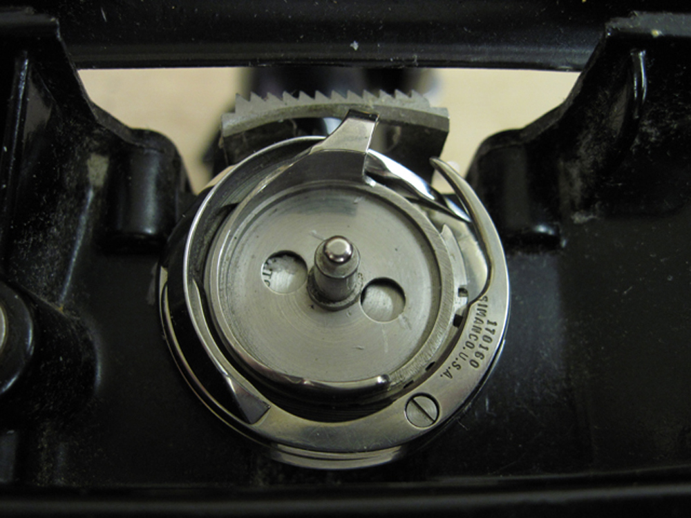 A closeup of the rotary hook mechanism, with the bobbin case and throat plate removed. The feed dogs are visible in the background.