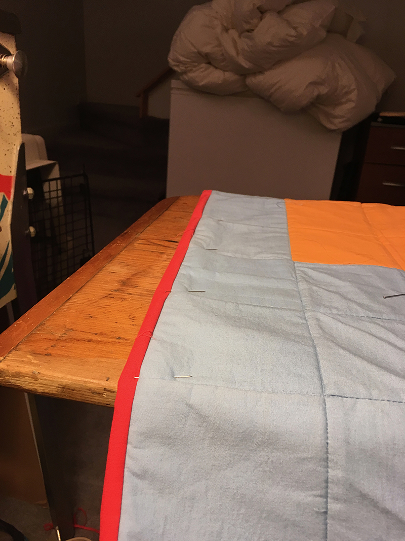 Red fabric binding tape is held in place at the edge of the quilt with dress pins.